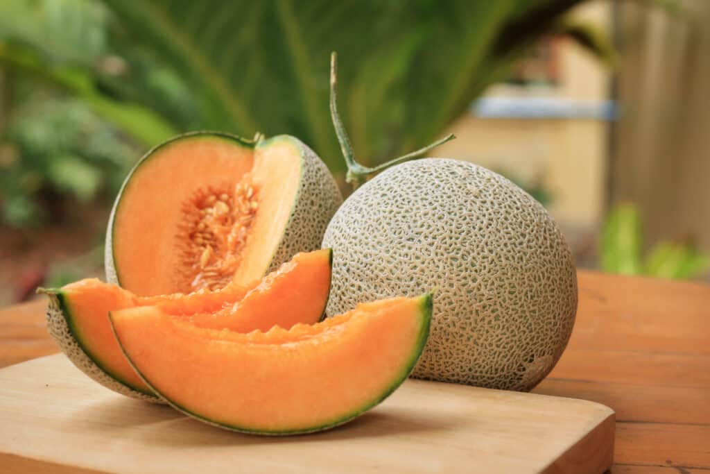fette di melone d'arancia su un tagliere con un intero melone sullo sfondo, insieme a un po' di verde.