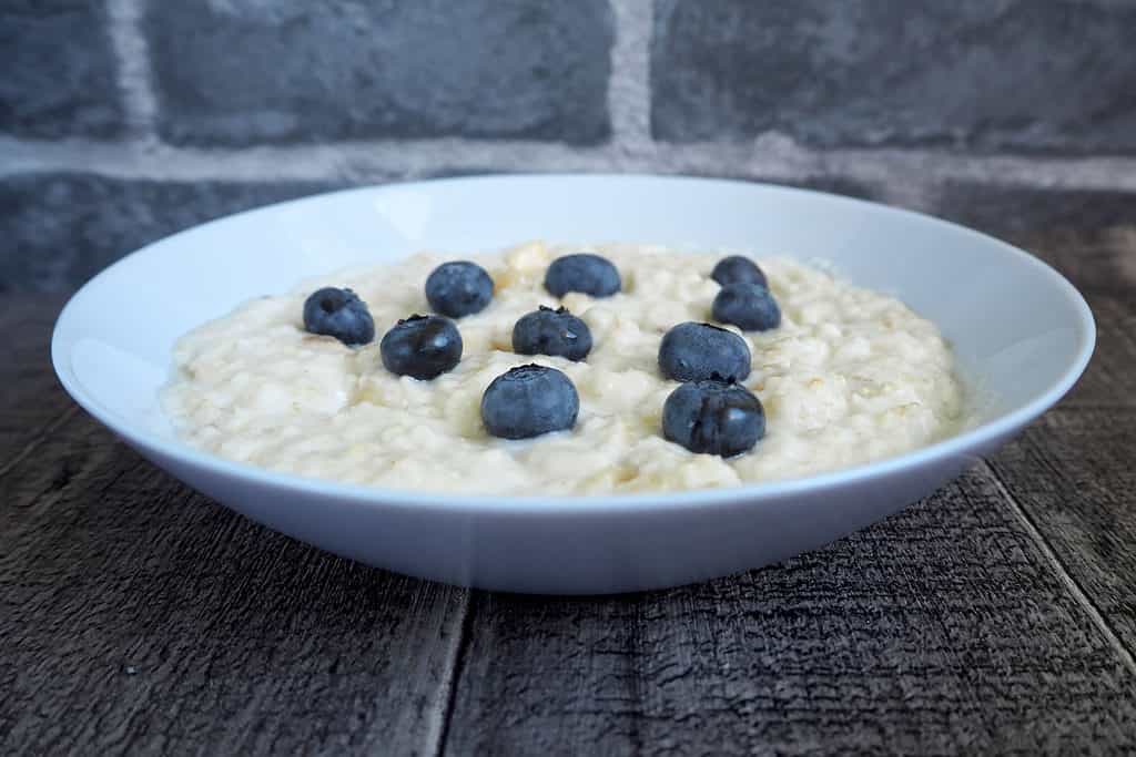 Ciotola di farina d'avena senza bordo in ceramica grigio chiaro, farina d'avena colorata, con 10 mirtilli mirtillo-blu disposti a triangolo, in 4 file dal riquadro sinistro.  la prima fila ha 1 mirtillo, la seconda fila ha 2 mirtilli, la terza fila ha 3 bacche blu e la quarta fila ha 4 mirtilli.  la ciotola è la cornice centrale di un tavolo di legno invecchiato/stagionato con un muro di mattoni grigio granito sfocato sullo sfondo