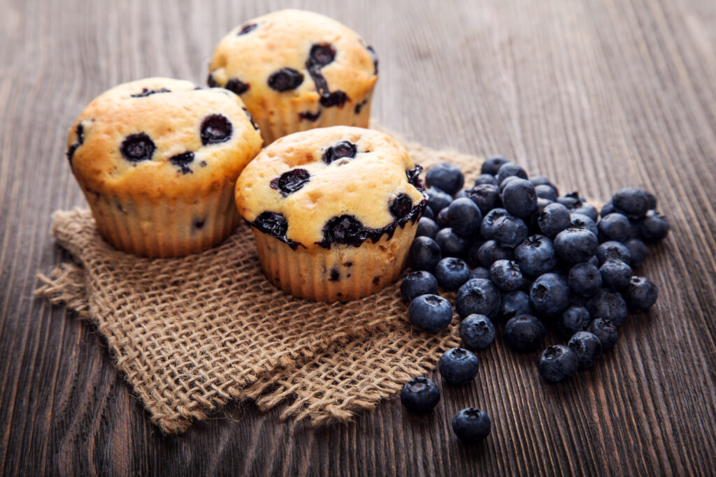 Cornice centrale: tre per lo più giallo oro con macchie blu, i muffin ai mirtilli sono stati posizionati esteticamente su un pezzo rettangolare di tela a trama larga, color tortora che è stato piegato a metà con una leggera angolazione. A destra dei muffin c'è una pila di circa 20 mirtilli, in parte sulla tela, in parte sulla tavola di legno che fa da sfondo. 