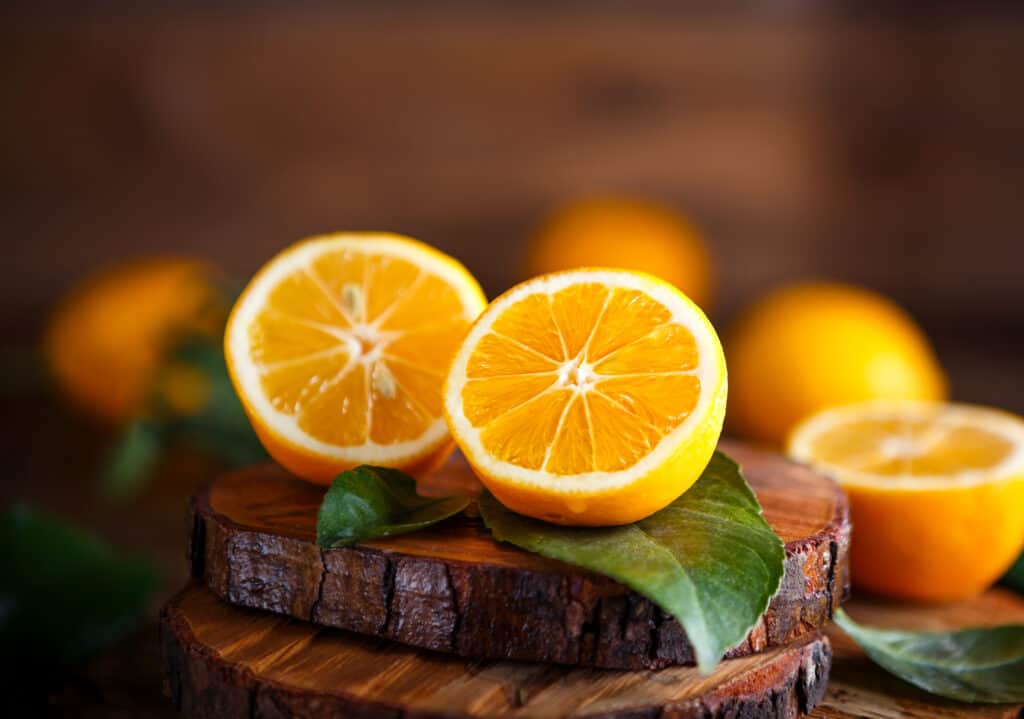 Un Meyer arancione tagliato a metà, seduto sopra foglie verdi, su una lastra naturale da 1/2 pollice di un tagliere di tronco di pino con un'altra metà di più due limoni Meyer arancioni interi. 