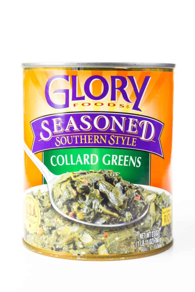 Can of Glory Foods Collard Greens condito in stile meridionale su sfondo bianco. L'etichetta della lattina è colorata. Il tardivo è arancione con riflessi verdi e viola. Nella parte inferiore dell'etichetta c'è una grande ciotola di verde cotto, con una grande cucchiaiata di verdure che copre sopra il centro della ciotola.