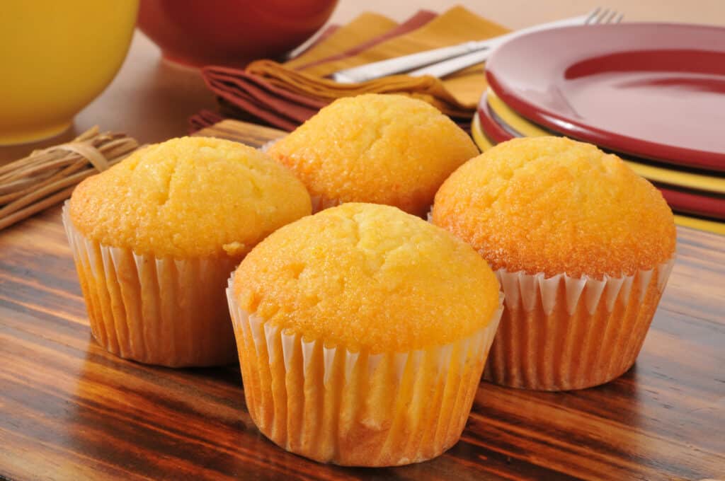Quattro muffin di pane di mais giallo dorato in carte cupcake di carta bianca raggruppate telaio centrale su un tavolo di impiallacciatura composita venatura del legno. dietro i piatti, in alto a destra, è visibile una pila di quattro piatti in gres rossi e gialli alternati visibili. a sinistra dei piatti si vedono alcuni tovaglioli gialli con forchetta e coltello in metallo argentato. Due grandi ciotole, davanti gialle, dietro rosse, sono in alto a sinistra accanto ai tovaglioli.
