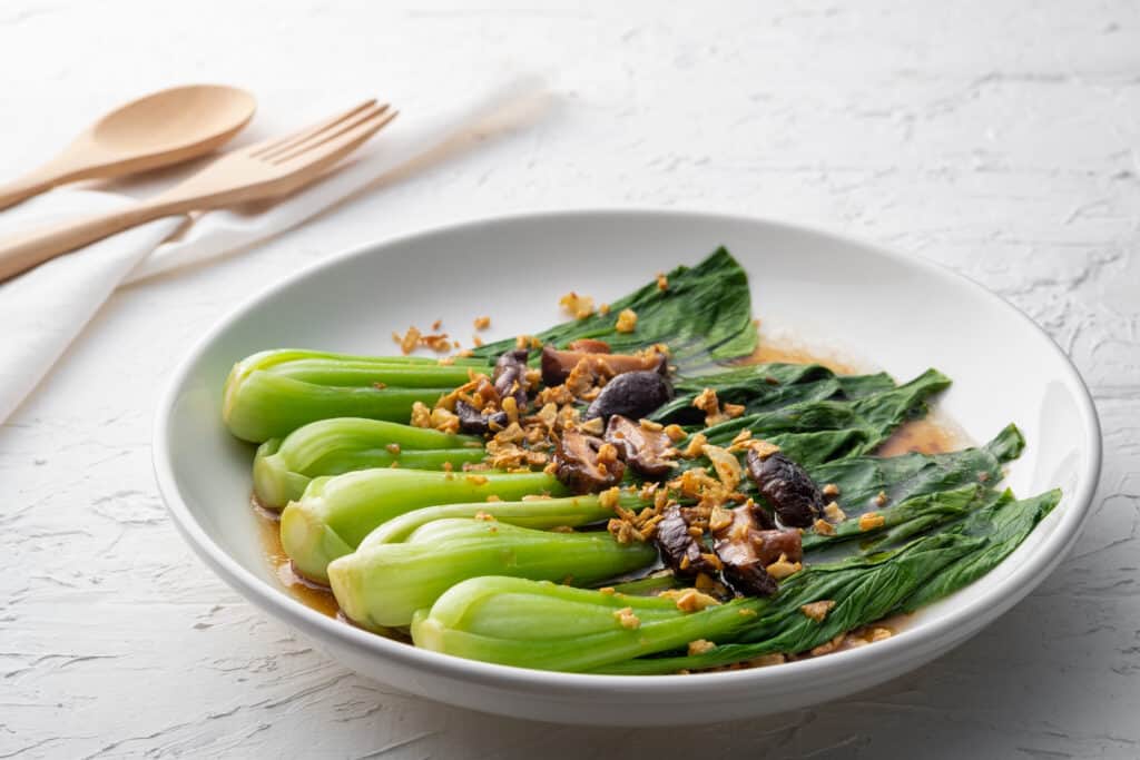 Una ciotola in ceramica bianca di baby bok choy in salsa di ostriche con funghi Shitake e aglio fritto.  Il cavolo cinese è cotto.  Le sue foglie sono di un verde più scuro dei suoi steli.  i funghi e l'aglio sono di diverse tonalità di marrone e oro.  una salsa/succo marrone è visibile nella parte inferiore della ciotola.  Sembra salsa di soia, ma è salsa di ostriche.  La ciotola si trova su un tavolo coperto da una tovaglia bianca.  un cucchiaio e una forchetta di legno (di bambù?) sono appoggiati su un tovagliolo dietro la ciotola.  Il tavolo coperto di stoffa fa da sfondo.