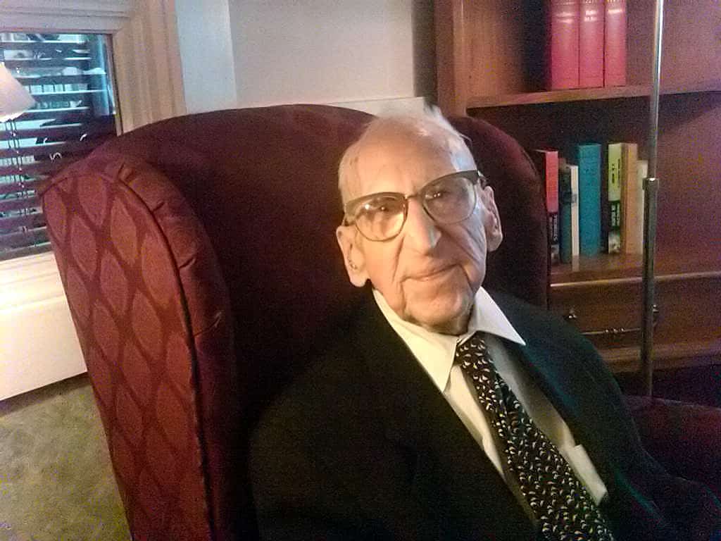 ritratto di Walter Breuning una volta l'uomo vivente più anziano del mondo.  Si sedette su una sedia imbottita rossa.  Indossa una giacca brome con una camicia chiara e una cravatta marrone.  Dietro di lui si vede una libreria piena di libri colorati.  La schiena della sedia YJe è rivolta verso una finestra/\.