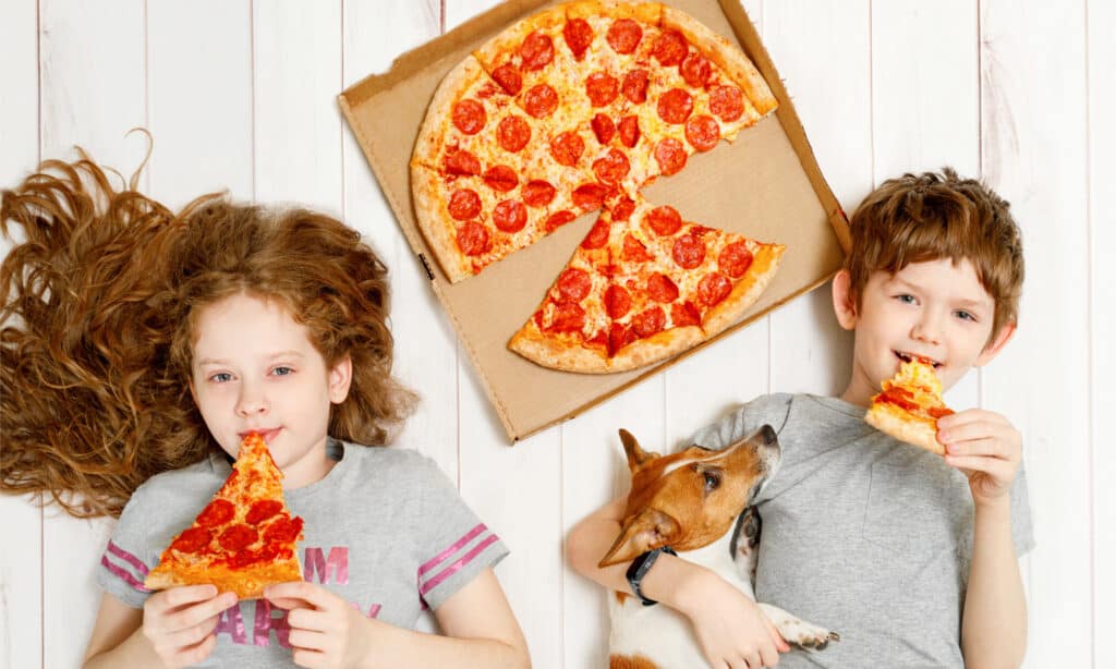 2 bambini sdraiati sul pavimento con il loro cane accanto a uno sfondo bianco di pizza ai peperoni