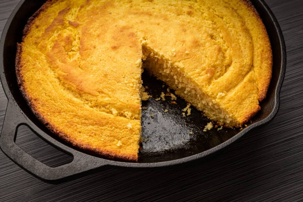 Una padella di ghisa con dentro del pane di mais dorato cotto. Manca un cuneo triangolare, tra le 4 e le 6. La padella è su un dannato tavolo di legno marrone.