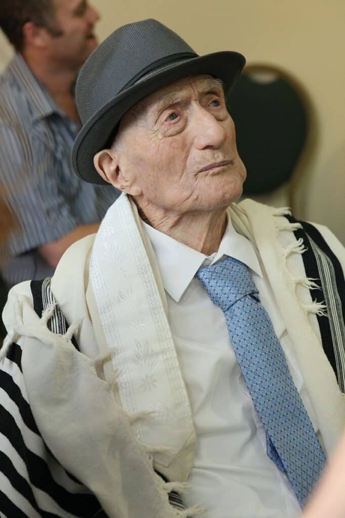 un ritratto di Yisrael Kristal, un tempo l'uomo vivente più anziano del mondo. Kristtal ha la pelle chiara, indossa un cappello fedora grigio scuro, uno scialle da preghiera, una camicia bianca e una cravatta azzurra.