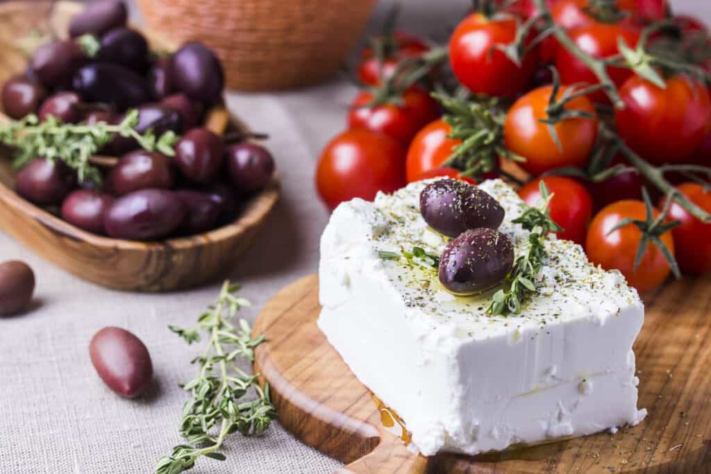 un rettangolo di feta greca su un tagliere di legno.  Il tagliere è su un tavolo coperto di stoffa chiara.  La feta ha sopra due olive viola/nere mature insieme a una spolverata di pepe nero macinato finemente e un rametto di timo.  Un altro rametto di tempo è sciolto sulla tavola, insieme a due olive mature.  Sullo sfondo (cornice a sinistra) c'è una ciotola di legno poco profonda ricolma di olive mature.  Dietro il formaggio (riquadro in alto a destra) ci sono una decina di pomodorini (ciliegini) arancioni, ancora attaccati alla vite, che sta seccando. 