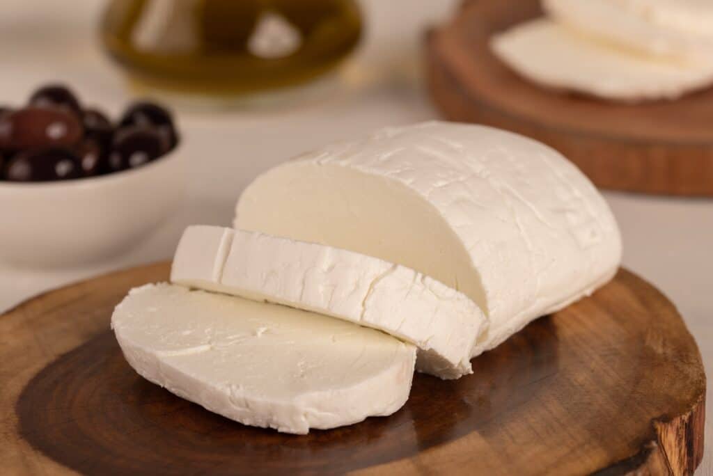 Cornice centrale: Karish Cheese.  un formaggio bianco morbido su una lastra di legno naturale, con due fette generose tagliate dalla massa principale di forma ovale.  Lo sfondo è costituito da olive nere mature in una ciotola di ceramica bianca, cornice a sinistra e cibo fuori fuoco nella parte superiore della cornice. 