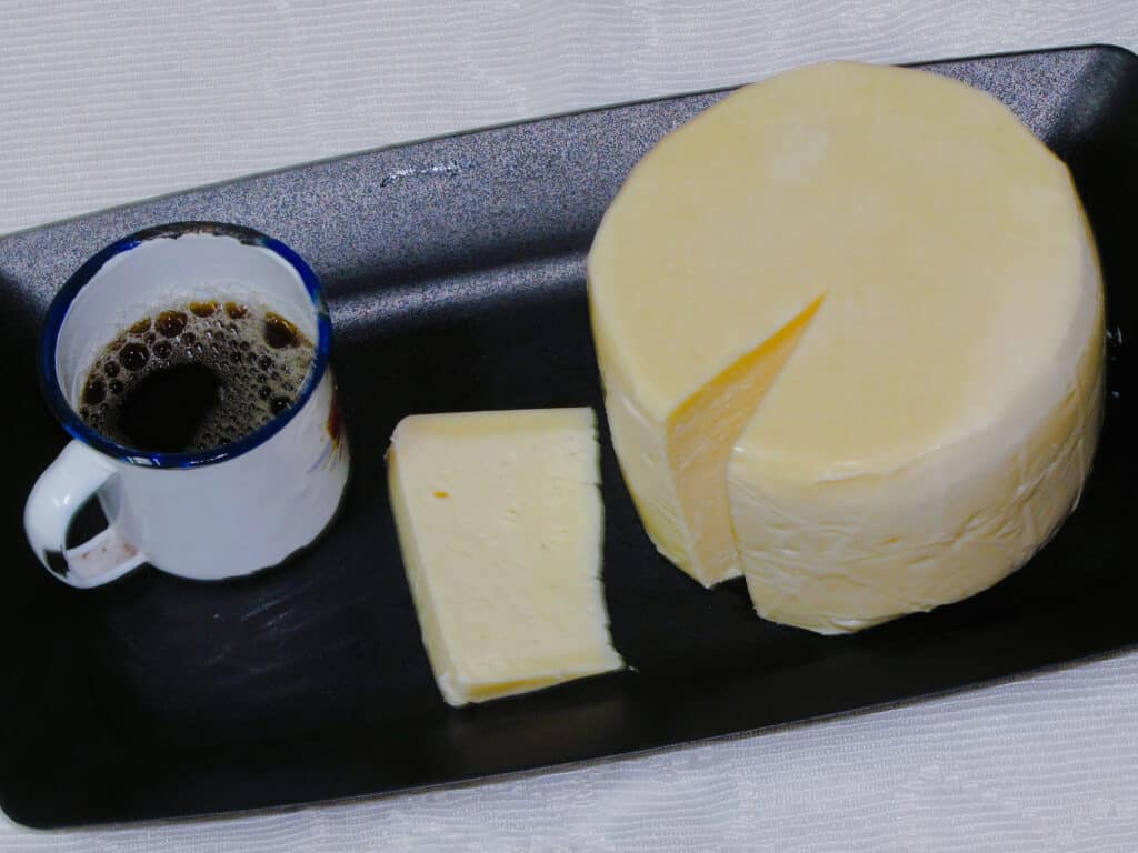 L'inquadratura è alta, guardando dall'alto in basso: una forma rotonda di formaggio Serro giallo molto chiaro - sembra burro.  Un cuneo sottile è stato rimosso e si trova accanto alla ruota/ Una tazza di metallo usata per ruota rivestita di smalto bianco e riempita per metà con un liquido marrone scuro con bolle marrone chiaro sulla sua superficie, si trova a sinistra della ruota e la fetta su un anonimo vassoio di metallo scuro.  contro uno sfondo bianco