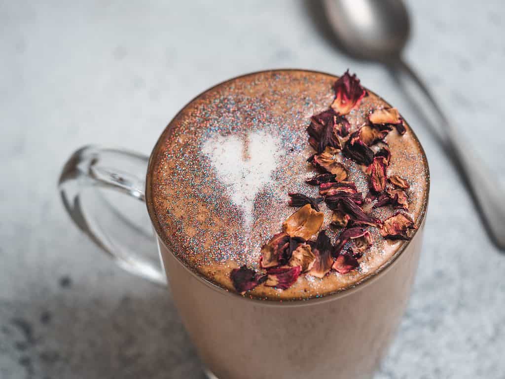 Caffè alla moda con glitter commestibili e petali di rosa essiccati.  Tazza di caffè scintillante o cappuccino diamantato sul tavolo grigio.