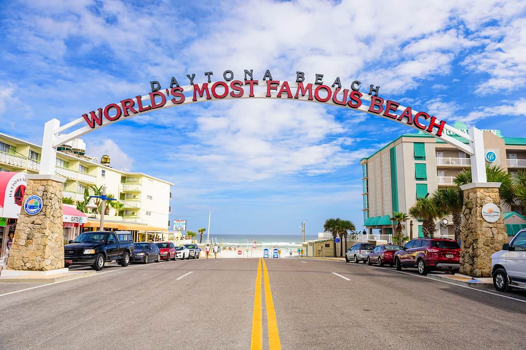 Daytona Beach, FL, USA - 2 febbraio 2015: I bagnanti in lontananza camminano sotto il cartello di benvenuto di Daytona Beach.  La popolare destinazione per le vacanze di primavera è soprannominata "La spiaggia più famosa del mondo."