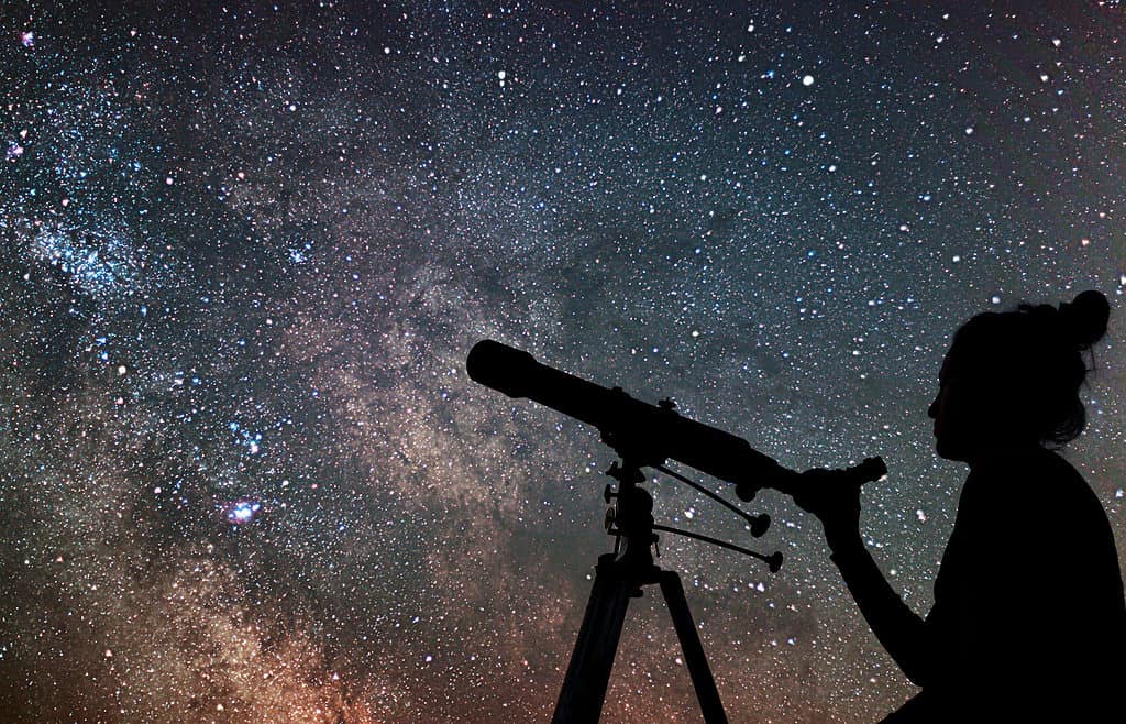 Foto cornice in basso a destra silhouette di una persona con i capelli raccolti in un nodo alto.  La mano destra della persona stringe un telescopio anche la sagoma, contro un cielo pieno di stelle