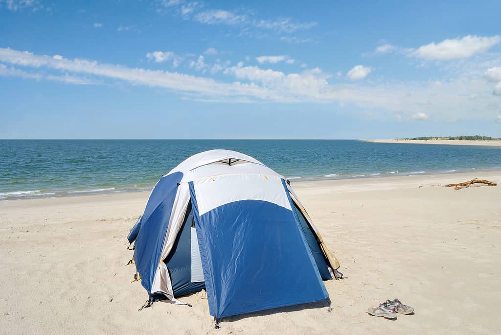 Foto di una tenda a cupola blu e argento su una spiaggia di un lago di sabbia bianca. C'è un paio di scarpe da corsa allacciate visibili vicino alla tenda. Il lago e un cielo azzurro con qualche nuvoletta bianca e sottile fanno da sfondo.