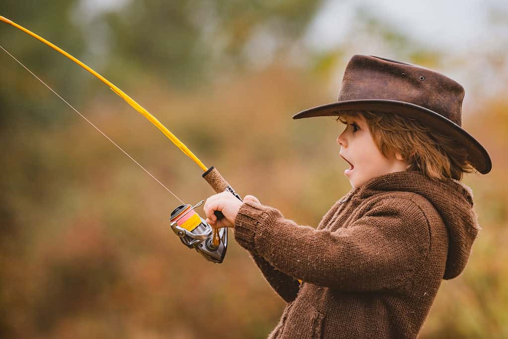Un bambino vestito con un maglione lavorato a maglia marrone a maniche lunghe.  Il bambino indossa un cappello a tesa larga in pelle.  Il bambino tiene in mano una canna e un mulinello gialli (canna da pesca).  Il bambino è rivolto verso l'inquadratura a sinistra e l'espressione sul viso sembra essere di euforia come se avessero appena pescato un grosso pesce.  Lo sfondo è fuori fuoco natura