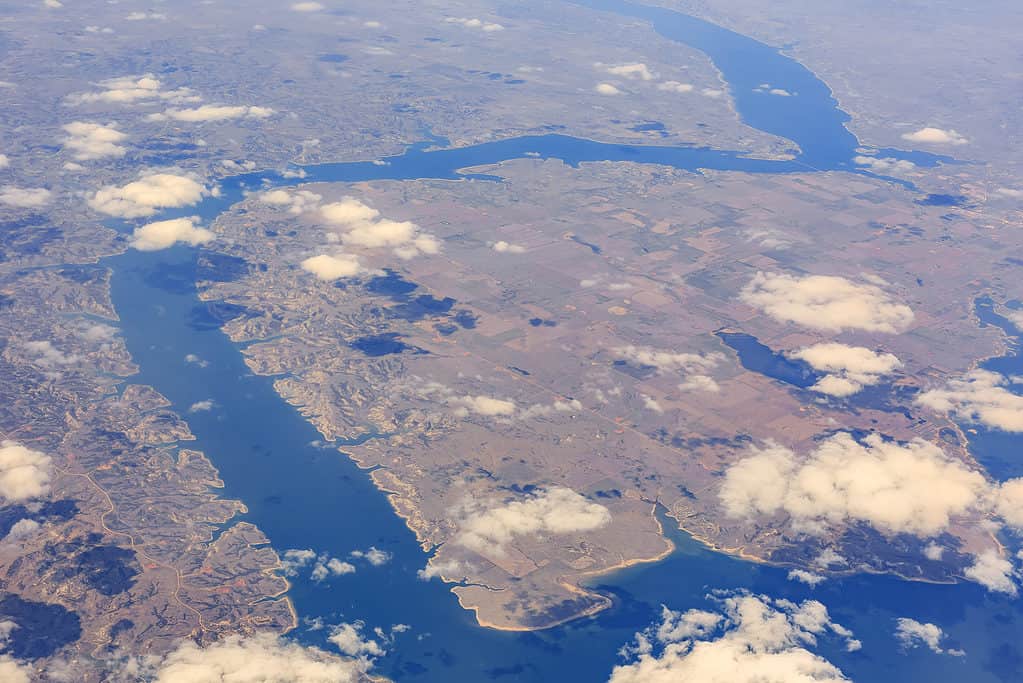 Una veduta aerea del lago Sakakawaea.  Il lago è stretto e si snoda attraverso l'inquadratura dall'inquadratura in basso a destra attraverso la parte sinistra della fotografia e poi serpeggia di nuovo a destra per circa 3/4 della salita e poi fuori dall'alto verso destra ci sono alcune nuvole tra la macchina fotografica e la fotografia e il treno è troppo lontano per sembrare qualcosa di diverso dal marrone e dal verde.