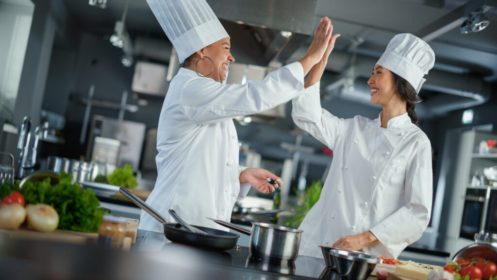Cucina del ristorante: Ritratto di donne chef asiatiche e nere che preparano il piatto, assaggiano il cibo, fanno il cinque in una celebrazione di successo.  Due professionisti che cucinano cibi deliziosi, autentici e sani