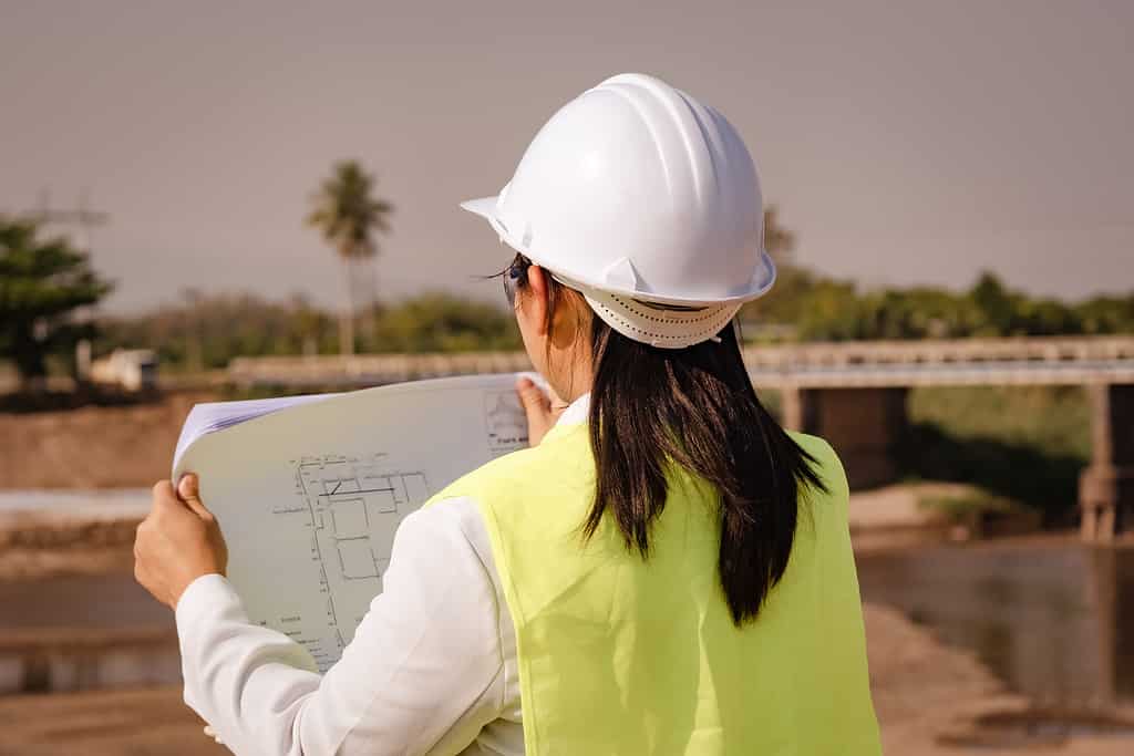 L'ingegnere che indossa un casco e un giubbotto di sicurezza lavora e guarda i progetti per il piano di costruzione e i dettagli di progettazione della diga.