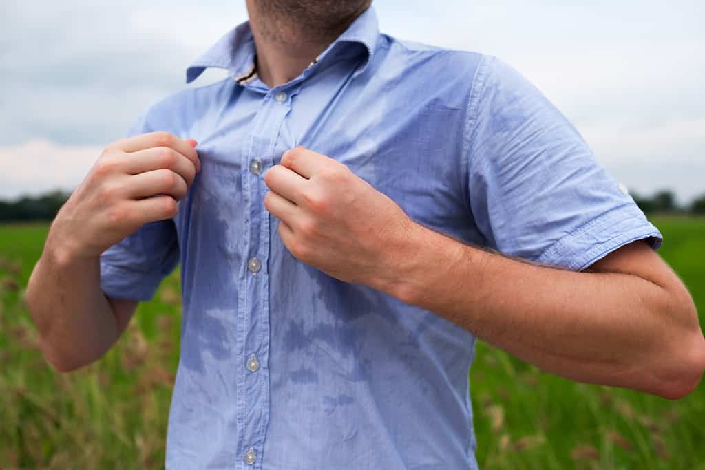 Uomo con iperidrosi che suda molto sotto l'ascella in camicia blu a causa del caldo.  Viaggiare in asia thailandia con zaino in spalla.  L'uomo si sta tirando la camicia, come per farsi vento.  La maglia è visibilmente bagnata di sudore.  L'uomo è abbronzato.  È in piedi in un campo.  Tutto ciò che è visibile è il suo mento e il busto.