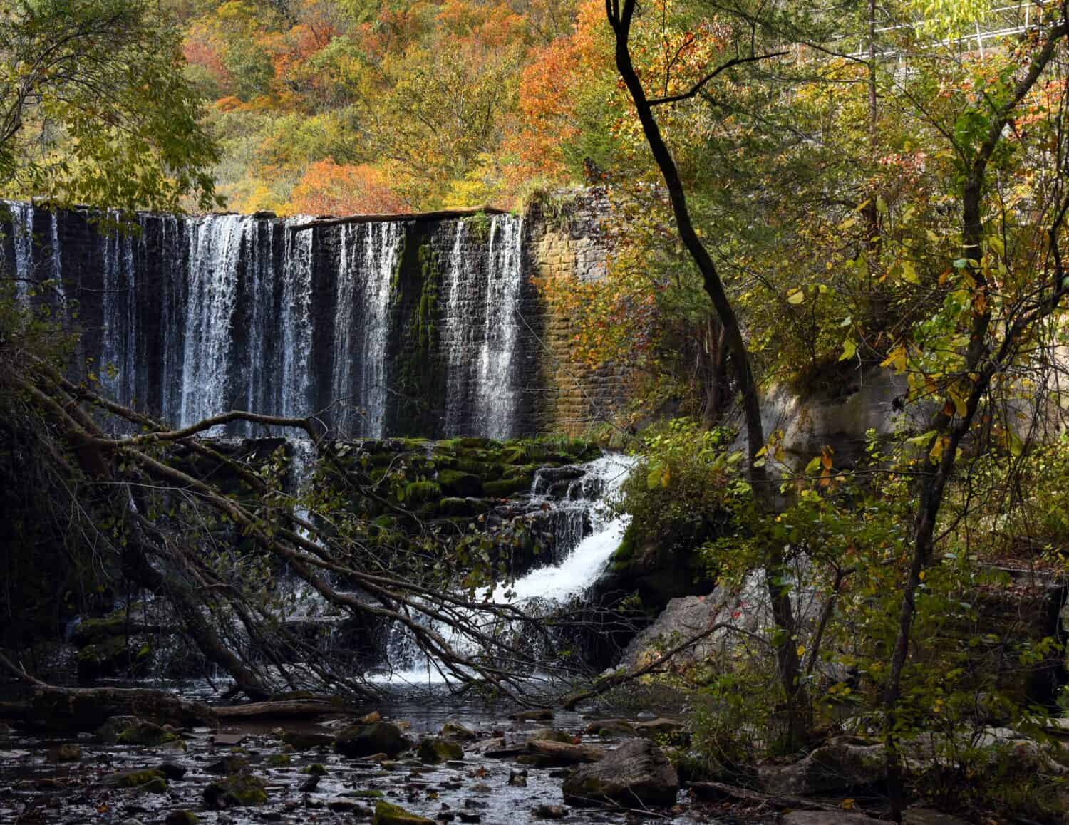 Vista della base della cascata di Mirror Lake. Lo sfioratore in pietra argina il lago ma consente all'acqua di fuoriuscire dalla diga. Le foglie autunnali riempiono il cielo nell'area ricreativa di Blanchard Springs Caverns.