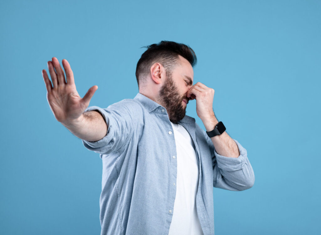 Concetto di cattivo odore.  Giovane ragazzo barbuto che chiude il naso e mostra il gesto di STOP, esprimendo disgusto sullo sfondo blu dello studio.  Uomo millenario respinto dall'odore tossico o dal profumo terribile