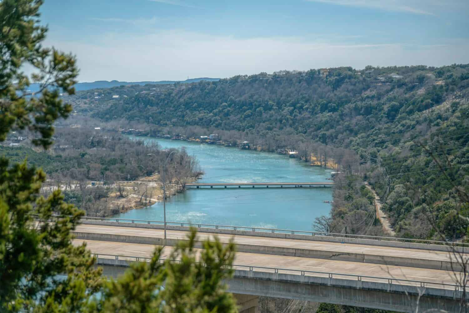 Vista dalla diga del lago Austin del fiume Colorado con ponte stradale in cemento.  Paesaggio aereo di acqua e carreggiata tra lussureggiante fogliame di alberi e cielo in una giornata di sole.