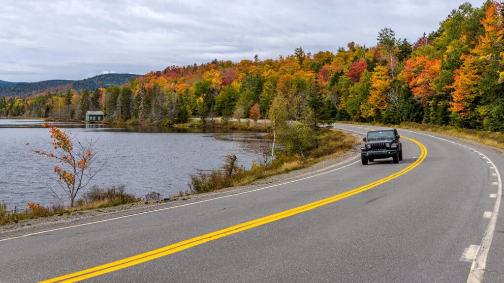 Jeep su Scenic Highway - Un SUV Jeep ultimo modello che guida lungo Beaver Pond sulla tortuosa Route 17, parte di Rangeley Lake Scenic Byway, in una mattina d'autunno nuvolosa ma colorata. Maine occidentale, Stati Uniti.