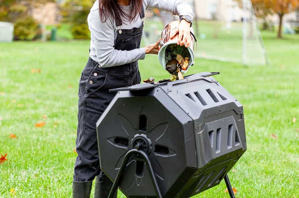 Una donna sta gettando un piccolo contenitore di scarti di cucina in un composter a rotazione esterna nel giardino sul retro. Queste unità di plastica con gambe in metallo possono girare per una migliore aerazione e un rapido compostaggio.
