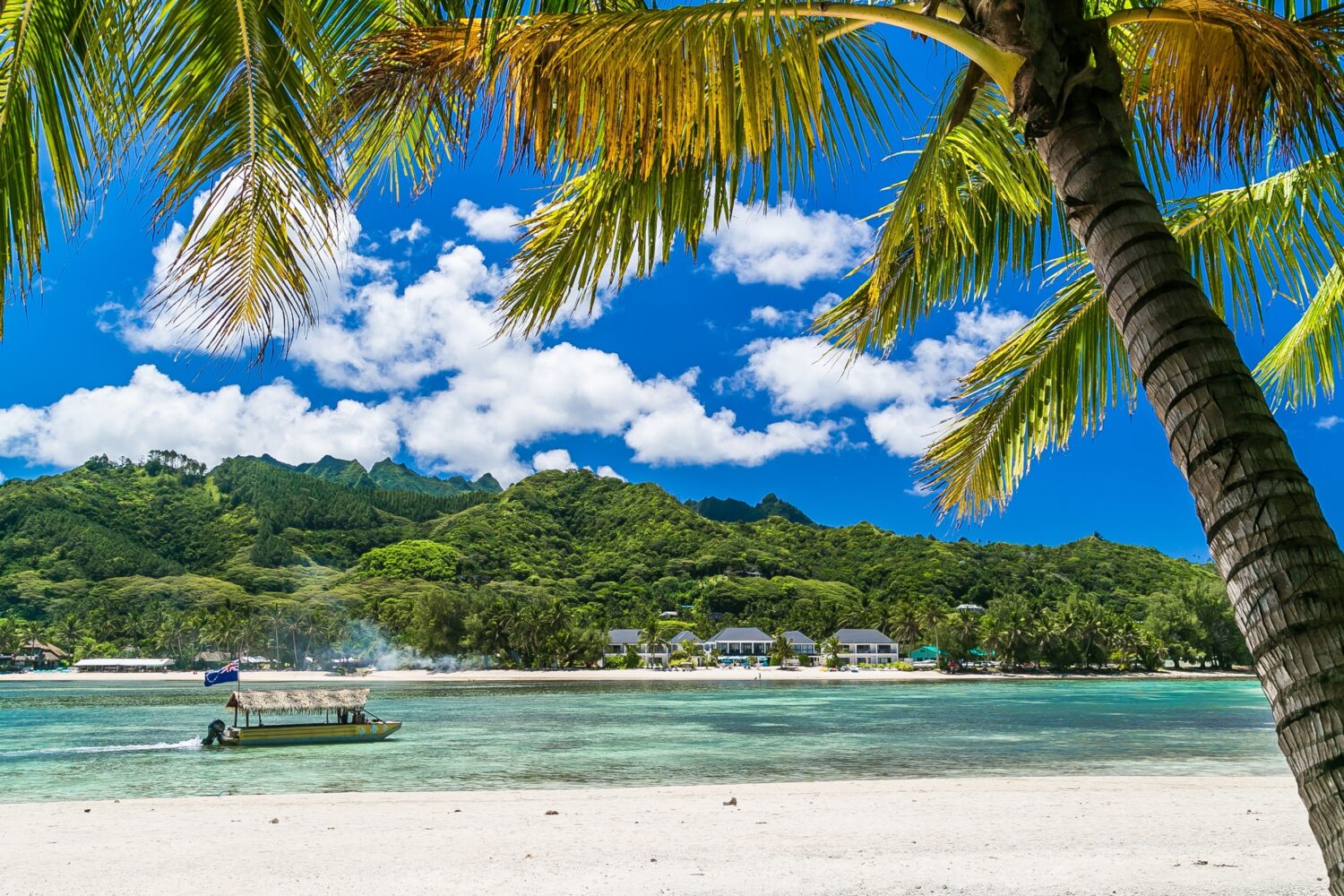 Baia tropicale, destinazione paradisiaca delle Isole Cook. Costa di Rarotonga con palme durante una giornata di sole. Una barca con la bandiera delle Isole Cook sul mare. Cielo azzurro con nuvole. Parzialmente nuvoloso.