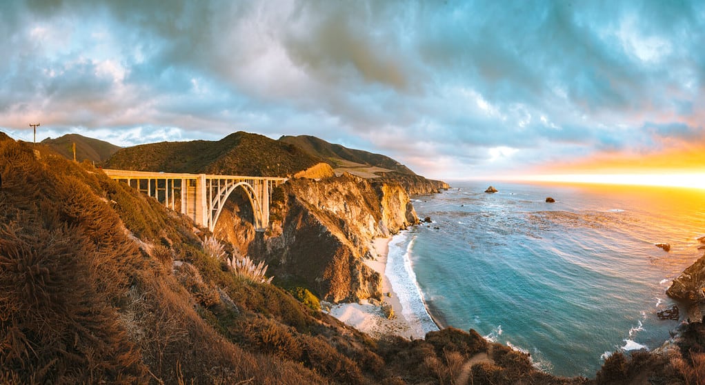 Vista panoramica panoramica dello storico ponte Bixby Creek lungo la famosa Highway 1 nella splendida luce dorata della sera al tramonto con un drammatico paesaggio nuvoloso in estate, Contea di Monterey, California, Stati Uniti