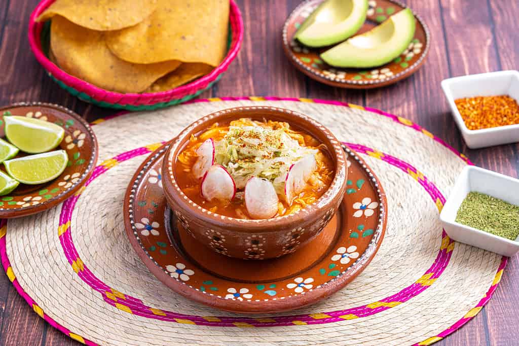 Cibo messicano tradizionale. Zuppa di pozole rossa con pollo accompagnata da avocado, lattuga, cipolla, ravanello, limone, peperoncino e tortillas di mais croccanti conosciute anche come tostadas su sfondo di legno.