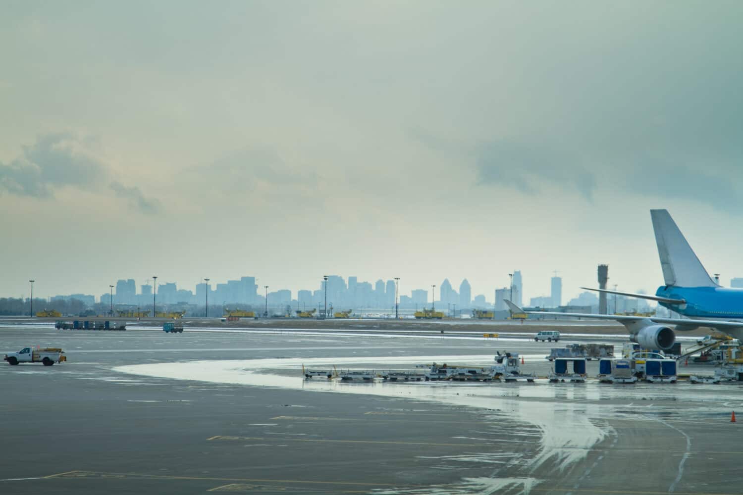 L'asfalto di un aeroporto, che mostra la parte posteriore di un aereo passeggeri e i veicoli di servizio che lo circondano, con lo skyline di una città dietro. Aeroporto Internazionale Pearson di Toronto, Ontario.