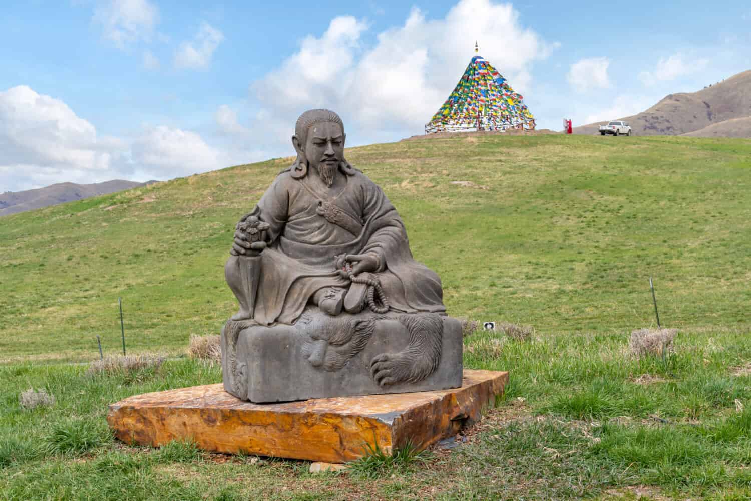 Una statua buddista di un guerriero o re seduto nella campagna rurale del Montana, con un teepee fatto di bandiere sfocate in lontananza.	