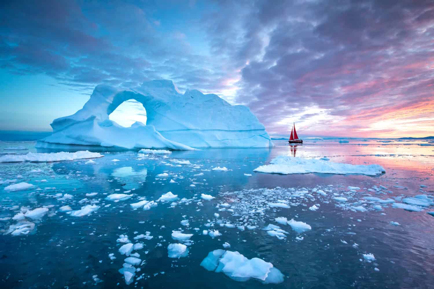 Piccola barca a vela rossa che naviga tra gli iceberg galleggianti nel ghiacciaio della Baia di Disko durante la stagione del sole di mezzanotte dell'estate polare.  Ilulissat, Groenlandia.
