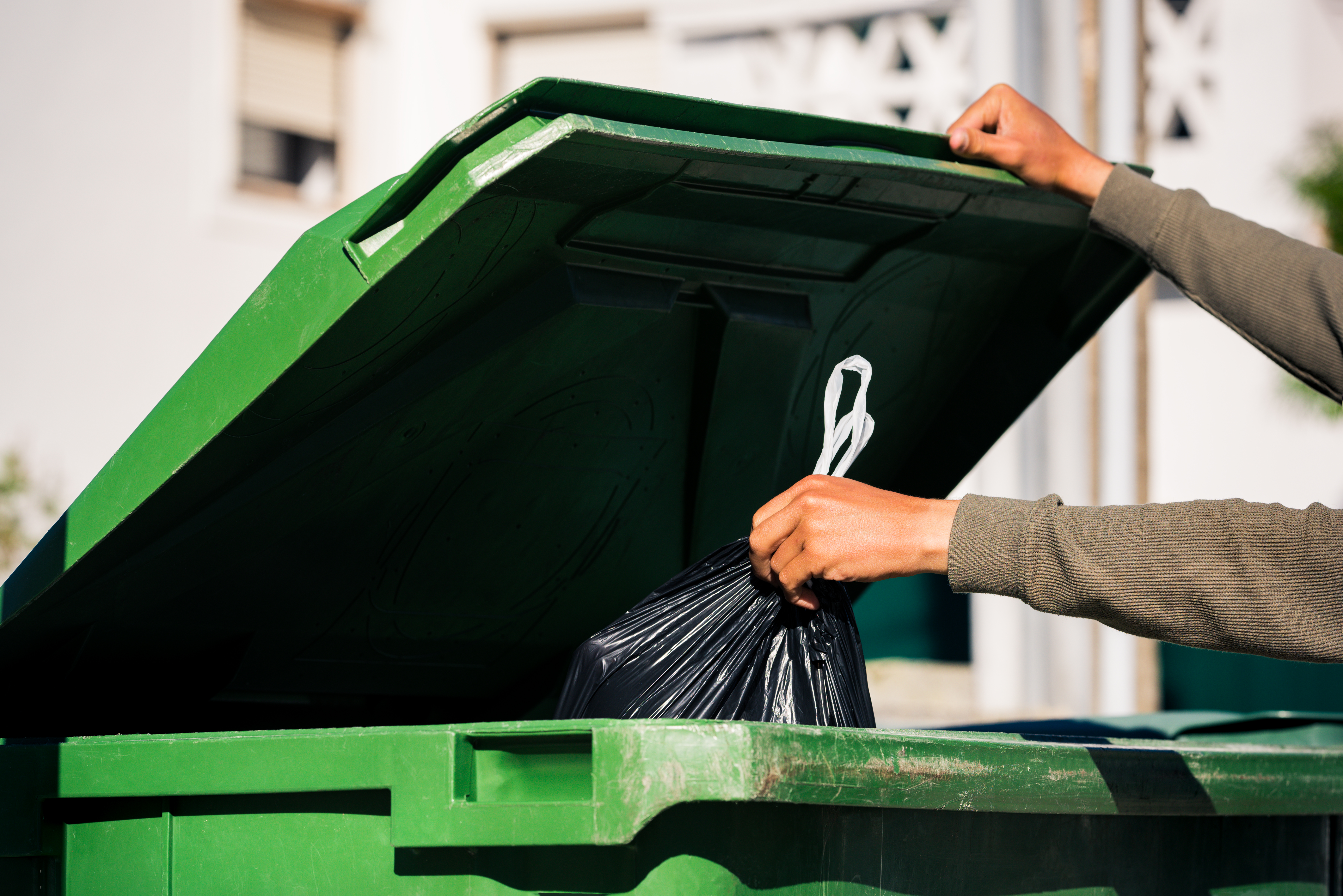 Uomo che butta fuori il sacchetto della spazzatura nero ecologico riciclabile in un grande contenitore per rifiuti verde in plastica. Porta fuori la spazzatura