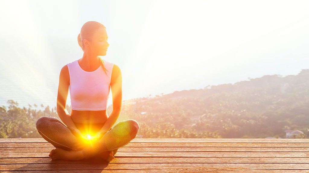 Bella donna seduta in una posa di mezzo loto in un luogo elevato con splendida vista dell'isola all'esterno, pratica la meditazione yoga illuminando il secondo chakra sacrale con gli occhi chiusi calmi.  Energia Kundalini