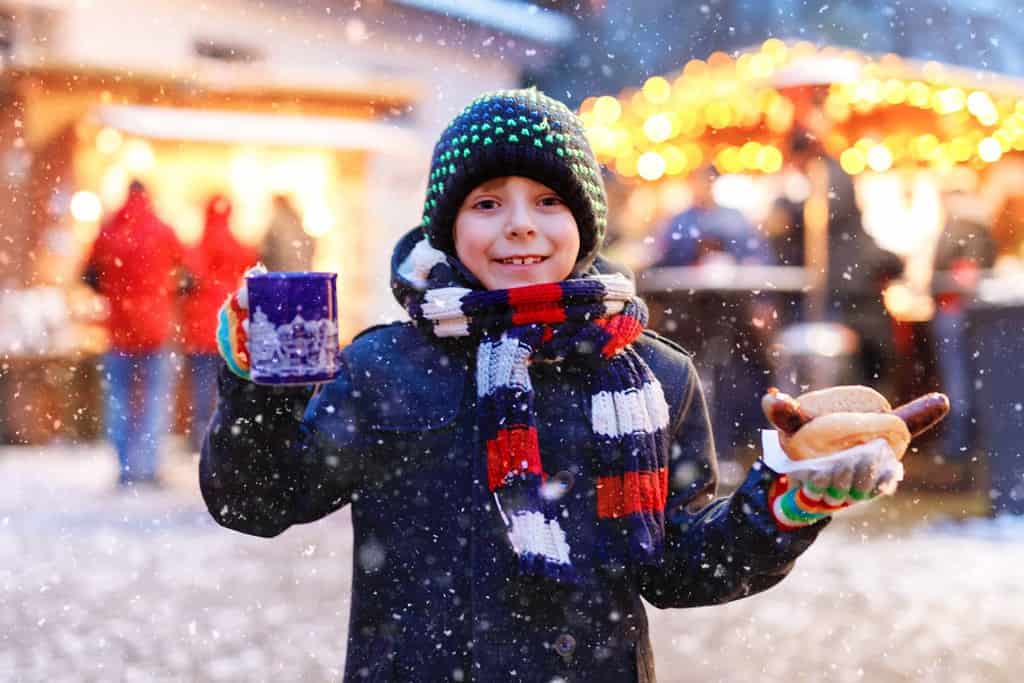 Un ragazzino carino che mangia salsiccia tedesca e beve un pugno caldo per bambini sul mercatino di Natale. Bambino felice sul tradizionale mercato familiare in Germania. Ragazzo che ride in abiti invernali colorati