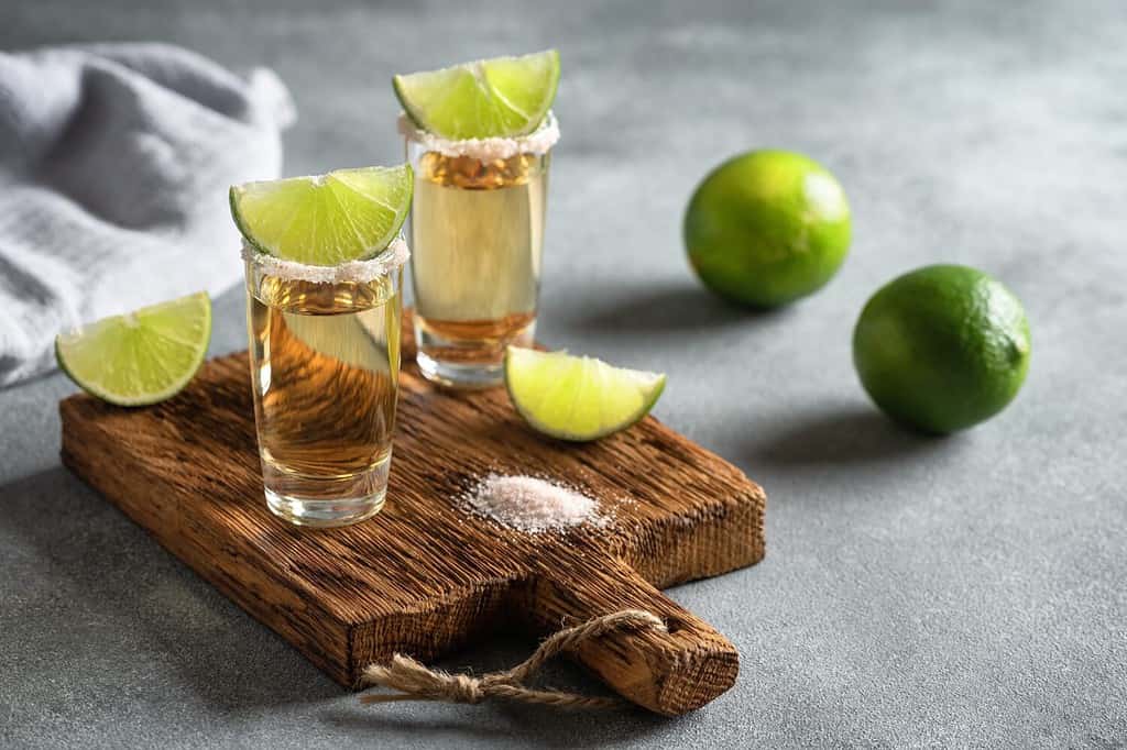 Tequila messicana d'oro in un bicchierino con lime e sale rosa su una tavola di legno, sfondo grigio cemento. Vista laterale, messa a fuoco selettiva.