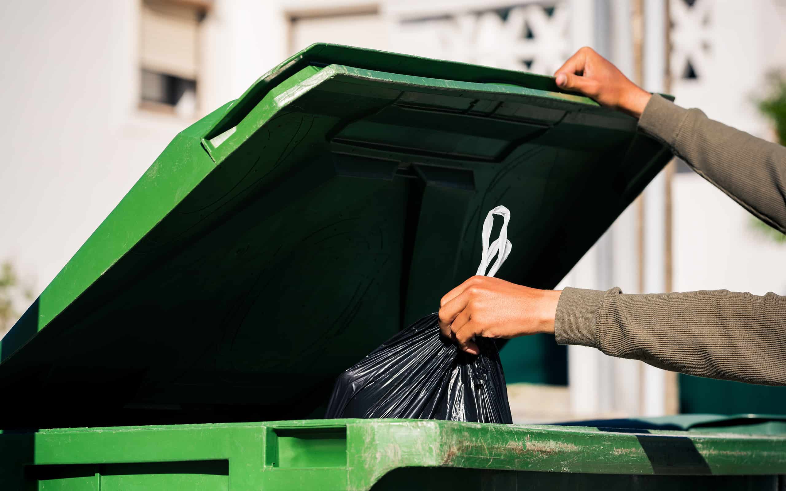 Uomo che butta fuori il sacchetto della spazzatura nero ecologico riciclabile in un grande contenitore per rifiuti verde in plastica. Porta fuori la spazzatura