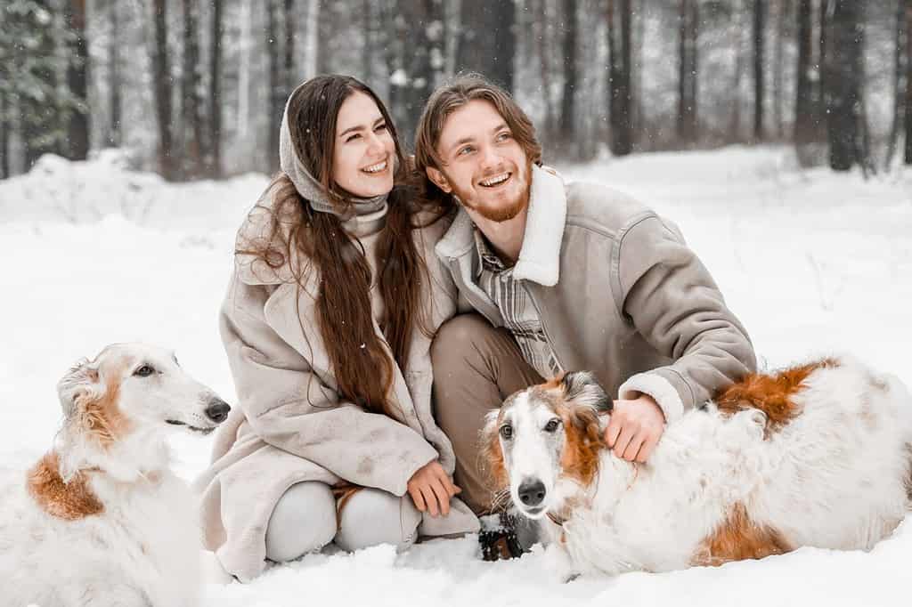 Adoro una giovane coppia romantica, un ragazzo nella foresta invernale fredda e nevosa che cammina con un animale domestico, un cane di razza borzoi russo da caccia. Levriero, proprietario di levrieri. Divertirsi, ridere. Pelliccia elegante, cappello di lana.