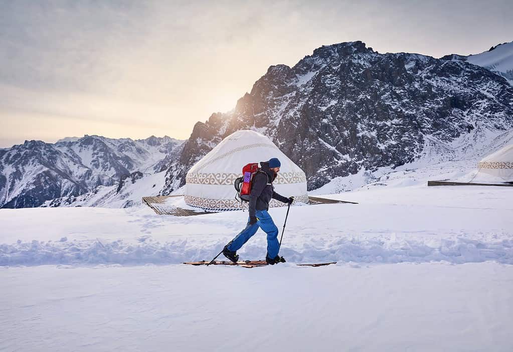 Sciatore uomo sci alpinismo in salita con barba e zaino in alta montagna innevata vicino alla tradizionale casa yurta nomade nella stazione sciistica Shymbulak ad Almaty, Kazakistan. Sport all'aria aperta in backcountry stagione invernale.