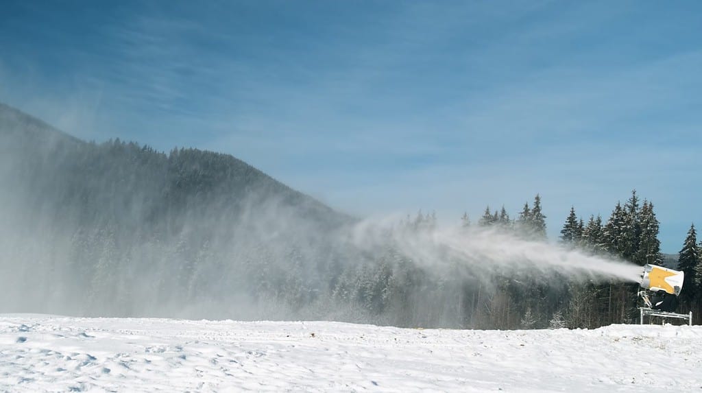 Il cannone da neve produce neve artificiale in una luminosa giornata di sole nella stazione sciistica. Sul retro foresta invernale, cielo blu. Gli impianti di innevamento spruzzano acqua per produrre neve. Preparazione della pista da sci per lo sci, gli sport invernali