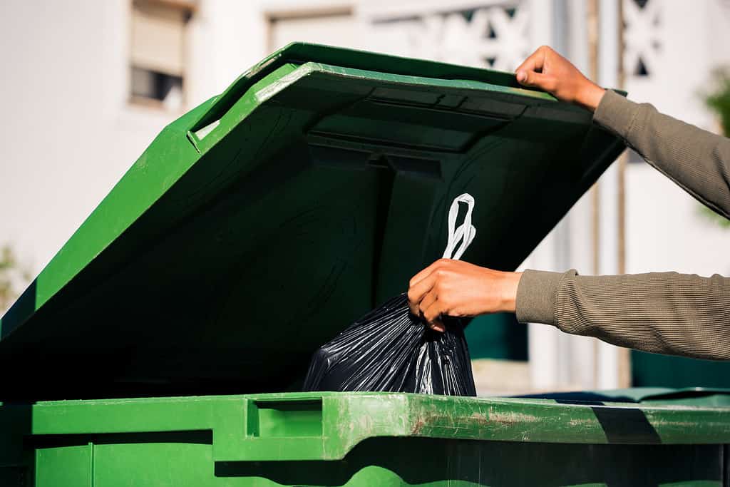 Uomo che butta fuori il sacchetto della spazzatura nero ecologico riciclabile in un grande contenitore per rifiuti verde in plastica. Porta fuori la spazzatura