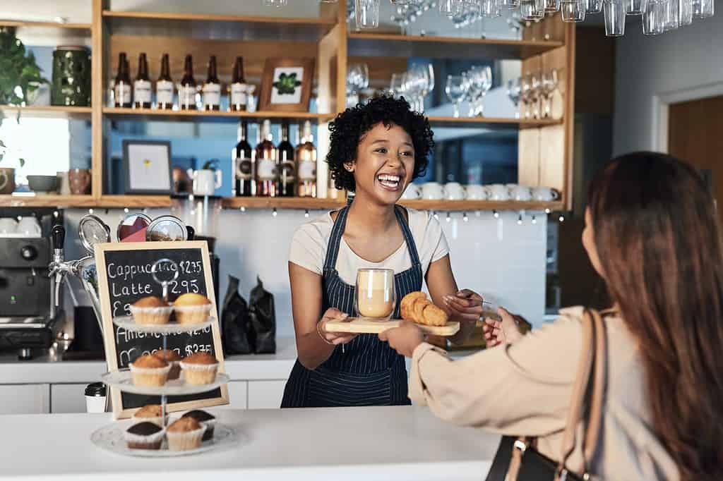 Donna felice, barista e cliente al servizio del bar per servizio, pagamento o ordine al bancone del bar. Persona africana, cameriera o dipendente in un ristorante di piccole imprese che aiuta il cliente alla cassa