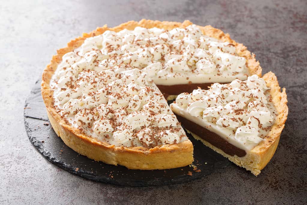 La torta Haupia al cioccolato è una torta cremosa con crema pasticcera al cocco ricoperta da una crosta di torta traballante e guarnita con panna montata in primo piano nel piatto. Orizzontale