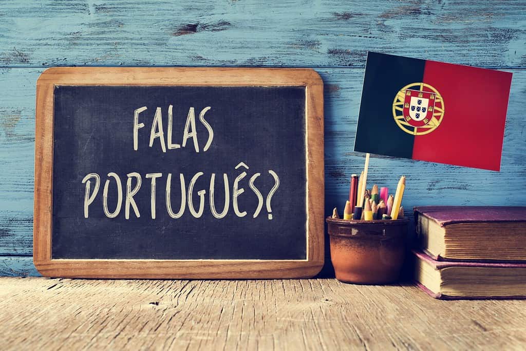 una lavagna con la domanda question falas portuges?  parli portoghese?  scritta in portoghese, un vaso con matite, alcuni libri e la bandiera del Portogallo, su una scrivania di legno