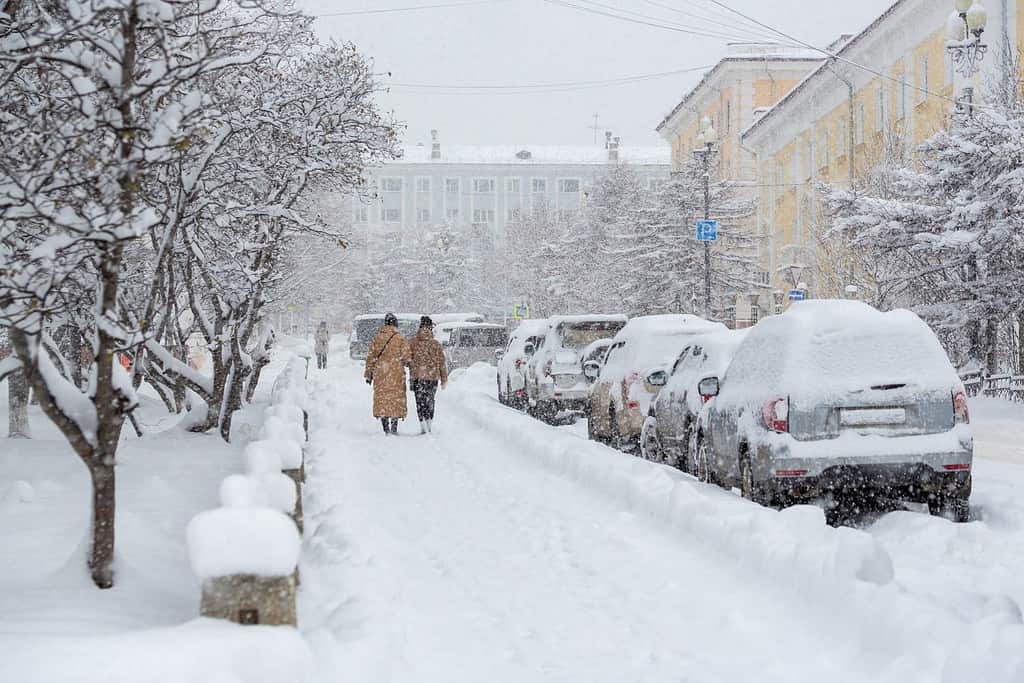Strada cittadina innevata durante una forte nevicata. Tanta neve sui marciapiedi, sulle auto e sui rami degli alberi. Le donne camminano per la città invernale. Tempo freddo e nevoso. Magadan, Siberia, Estremo Oriente russo.