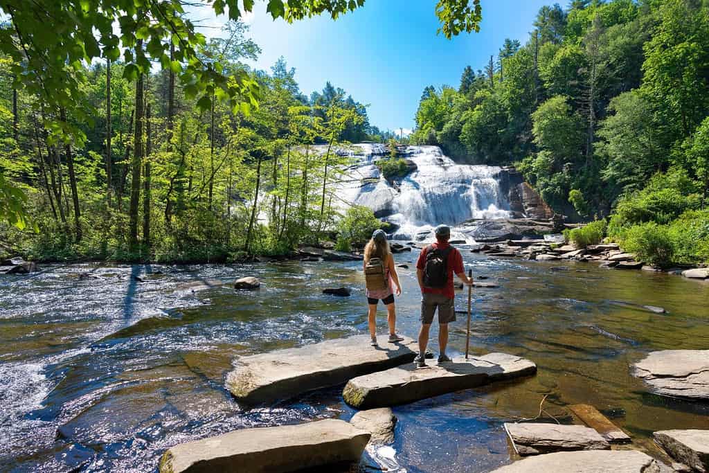 Coppia in piedi sulla roccia godendo della splendida vista sulla cascata. Amici che si rilassano durante un'escursione. Alte cascate della foresta demaniale di Dupont a Brevard.  Montagne Blue Ridge, vicino ad Asheville, Carolina del Nord, Stati Uniti.